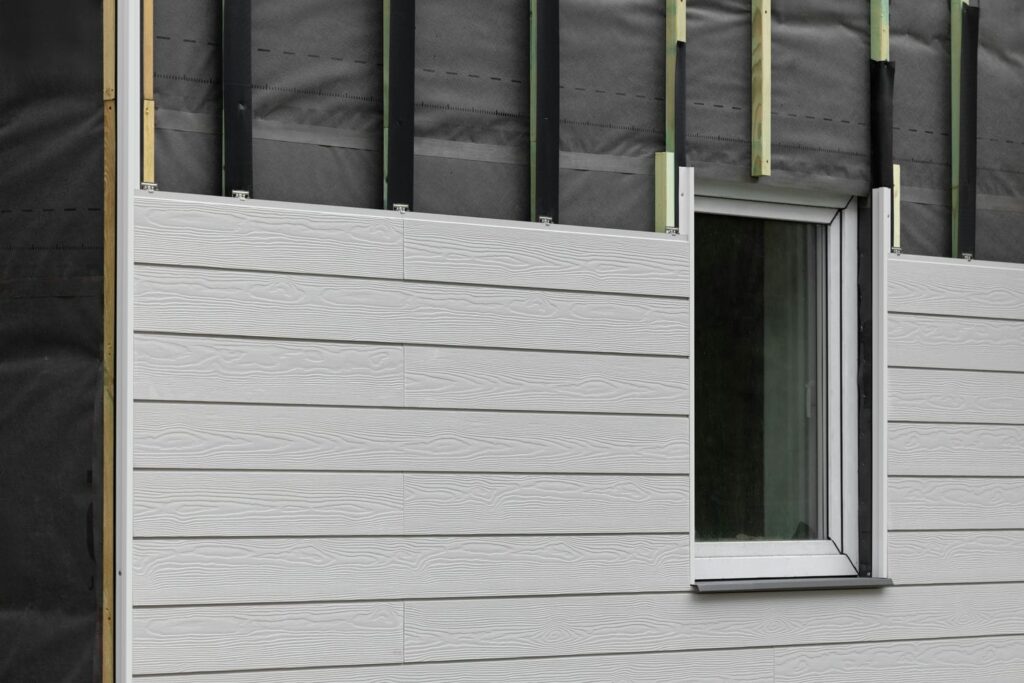 Fiber cement siding being installed over rain screen strapping and weather barrier next to a window opening.